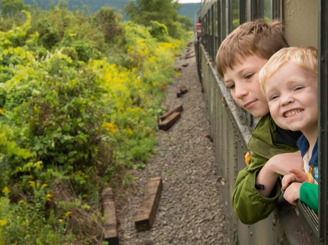 Two children leaning out of a train window with plant life to the left of the track.