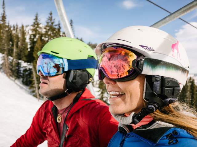 A man and a woman wearing helmets and goggles riding the ski lift at Salt Lake's ski resorts