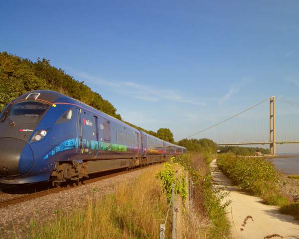 Blue Hull Trains trains speeding past the Humber Bridge (towards the camera) on a sunny day.
