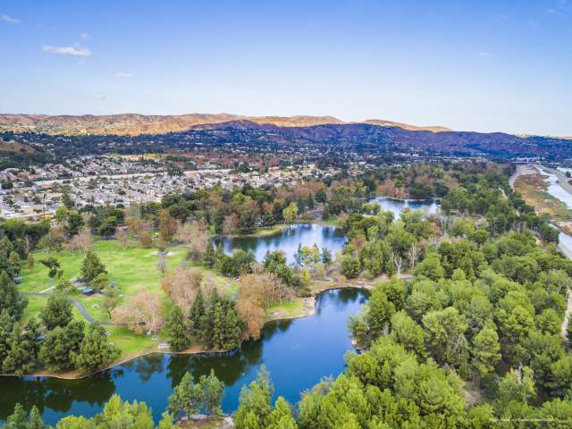Yorba Regional Park in Anaheim featuring an aerial view of tree-lined lakes, winding walking paths, open green spaces, and surrounding residential neighborhoods under a clear blue sky.