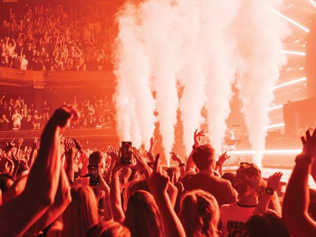 Photo from the audience looking up at the stage at ACL Live music venue. There are smoke machines at the front, and the room is tinged in red/orange light