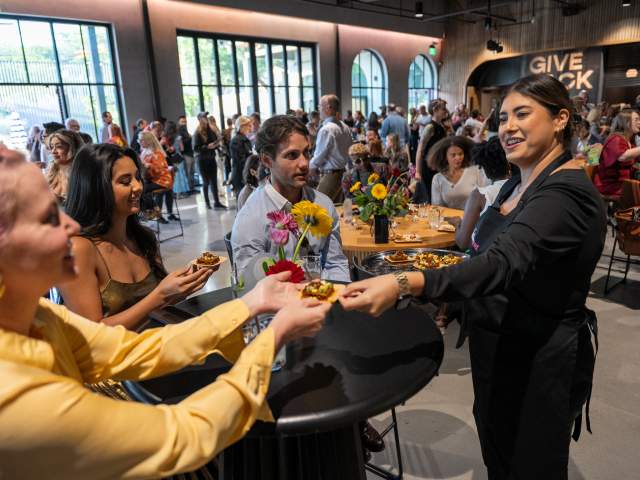 Student chef handing a bite of food to someone at a table of Give Back Gig attendees.