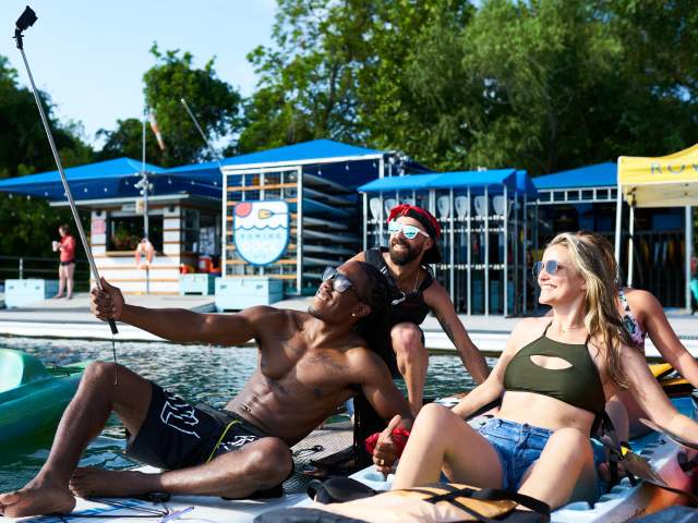 Photo of four people sitting on rental paddle boards in front of the Rowing Dock rental shop on Lady Bird Lake. They are all smiling and looking towards a camera on a selfie stick