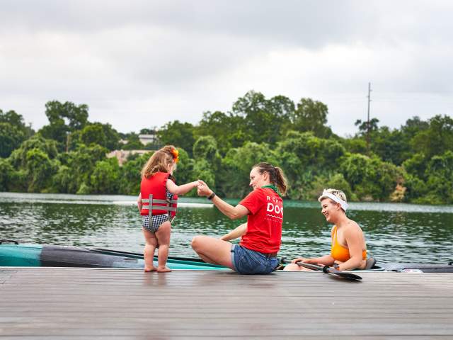 Photo looking towards the back of a child in a red life vest, she is standing at the edge of a dock on Lady Bird Lake and holding the hand of a woman in a Rowing Dock tshirt. Another woman is sitting in front of them in a rental kayak smiling at the young girl