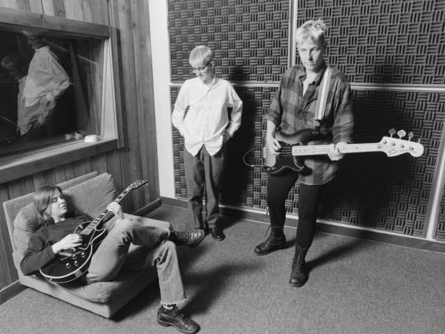 Photo of Meg Hentges and Darcee Douglas and another musician in a studio Douglas is standing on the right and playing the guitar, Hentges is standing in the middle and looking down and a third musician is sitting on a cushion on the left and playing the guitar