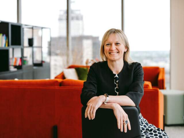 Katie Trout dressed in black jacket, sat on black chair in front on orange sofa in an office with Birmingham skyline in the background