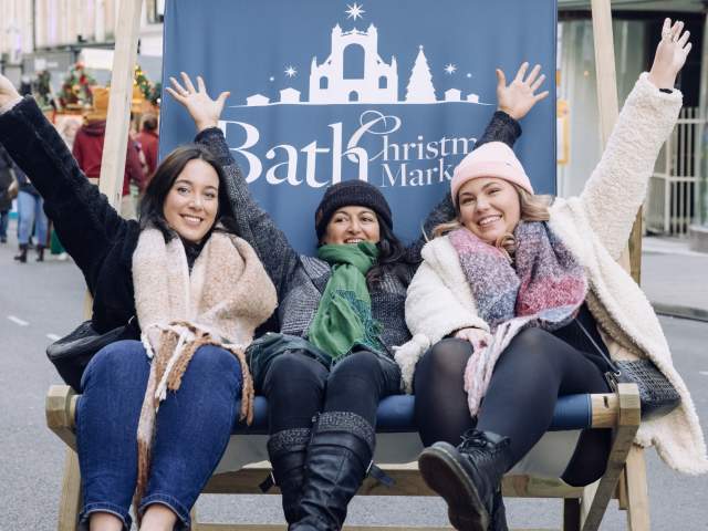 3 women sat in a big blue beach style chair that says Bath Christmas Market