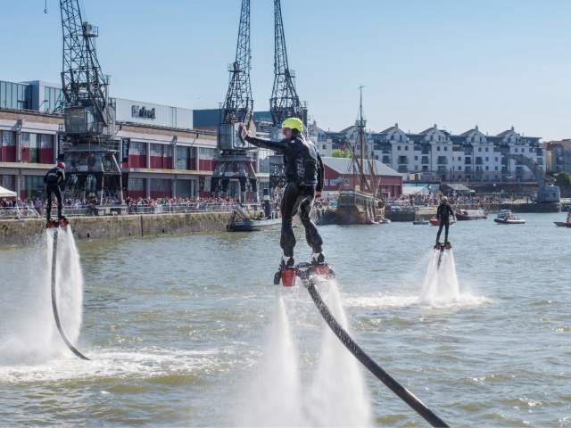 A man wearing a water-powered jetpack performing stunts on Bristol's Harbourside during the Bristol Harbour Festival with the M Shed local history museum in the background - credit Jim Cossey