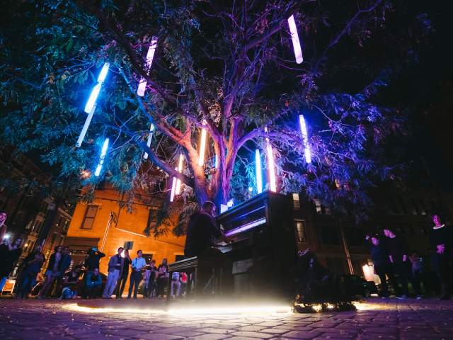 A man plays piano under a tree adorned with lights while others watch.