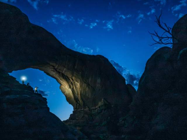 A group of night photographers standing beneath the towering span of Double Arch in Arches National Park near Moab, Utah.