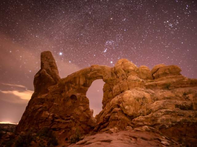 Turret Arch glowing against a star-filled night sky with the Orion constellation in Arches National Park near Moab, Utah.