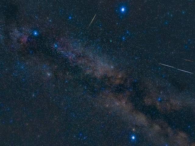 A deep-sky photograph highlights the three brilliant anchor stars of the Summer Triangle asterism framing the Milky Way, accompanied by several streaking Perseid meteors.