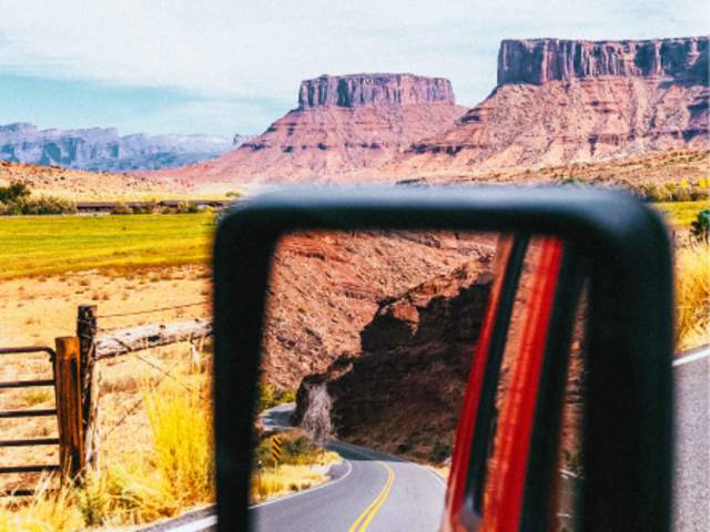 A side-mirror shot captures the dramatic red rock landscape along Utah State Route 128 (SR-128), commonly referred to by locals as the Moab River Road or the Upper Colorado River Scenic Byway.