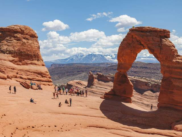 Delicate Arch pictured with a group of about 15 people looking at it. In the background, the La Sal Mountains are topped with snow.