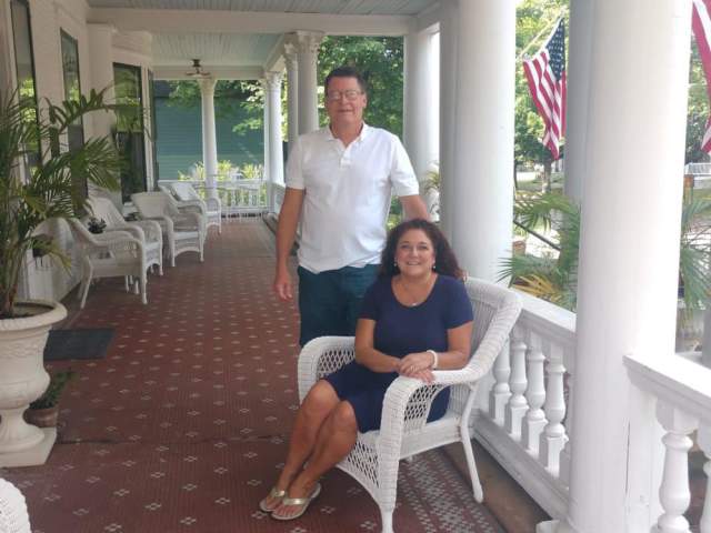 two figures pose and smile on a colonial porch