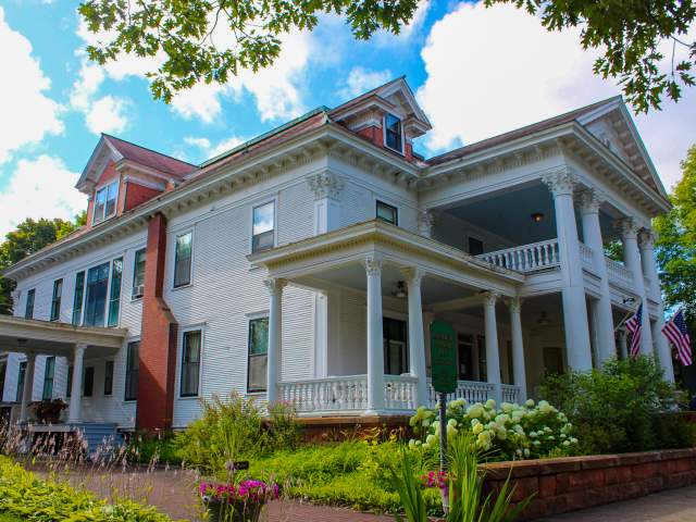 an antebellum style mansion with a wrap around porch is highlighted by blue skies and lush summer foliage