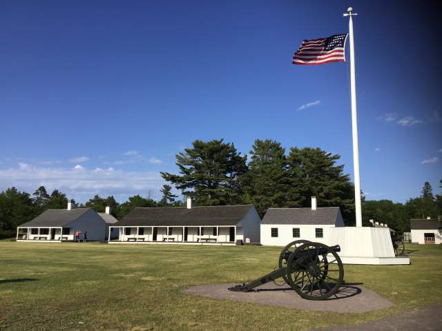 a historic canon sits on an open lawn, historic buildings and the American flag in the background