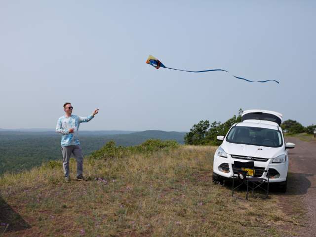 a person smiles as they stand near a cliffside while flying a kite