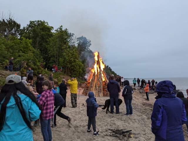 a crowd gathers around an enormous beachside bonfire