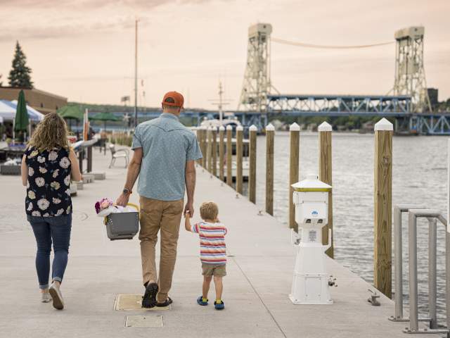 two adults guide a toddler on a waterside pier alongside a farmers market