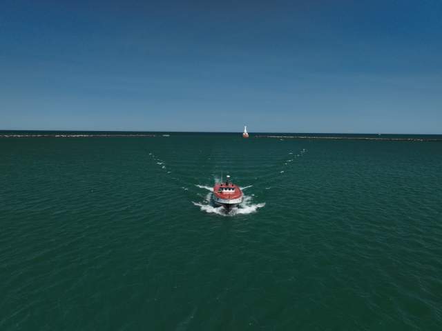 a boat on open water beelines toward the camera, a lighthouse in the distance
