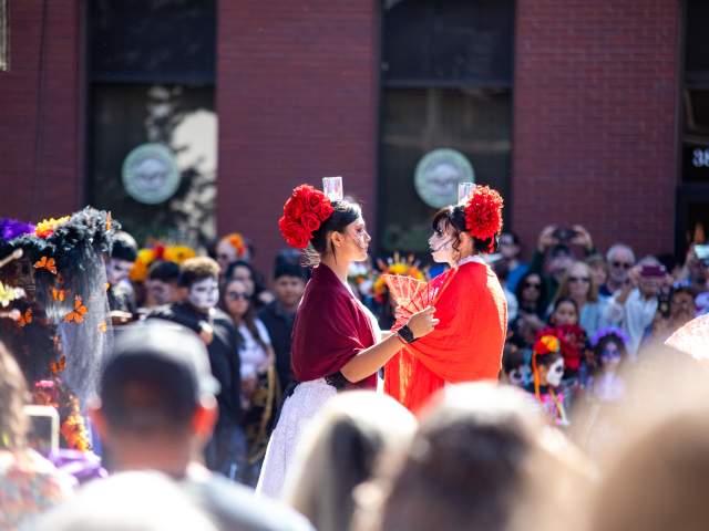 Two women performing in bright colors at the Longmont Day of the Dead festival with a crowd surrounding.