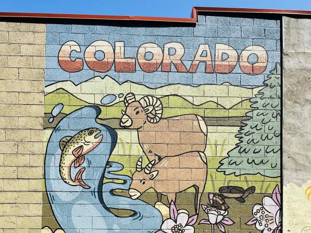 A mural on a brick wall that has images of two rams, a fish splashing up in water, flowers, trees, and a mountainous green background