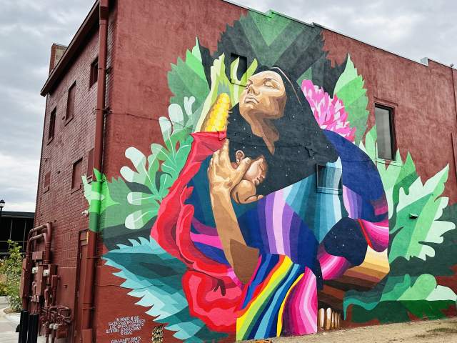 A mural on a downtown dark red building of a woman with dark hair holding a child to her chest and n ear of corn on her back with foliage surrounding them.