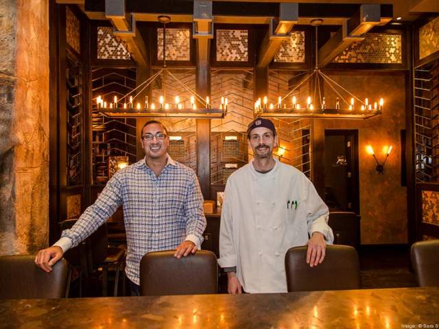 Two men stand behind chairs in a warm, dimly lit restaurant. One wears a plaid shirt and glasses, the other is dressed in a chef’s coat and hat. Behind them are decorative wine racks and two large chandeliers with glowing candles.
