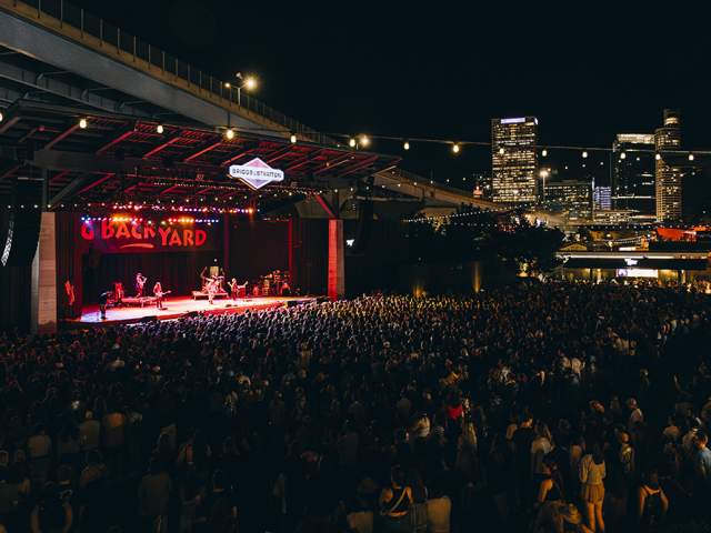 A large crowd gathers at night under a highway overpass for a live outdoor concert at the Briggs & Stratton Big Backyard stage at Summerfest in Milwaukee. The stage is lit with vibrant red and yellow lights as a band performs. String lights hang above the crowd, and the downtown Milwaukee skyline glows in the background.