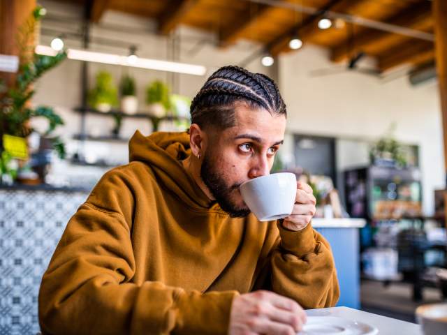 A man with braided cornrows and a trimmed beard sits at a table in a modern coffee shop, sipping from a white mug. He is wearing a brown hoodie and looking off to the side with a focused, thoughtful expression. The background is softly blurred, showing warm wooden ceiling beams, hanging lights, and green plants on shelves.