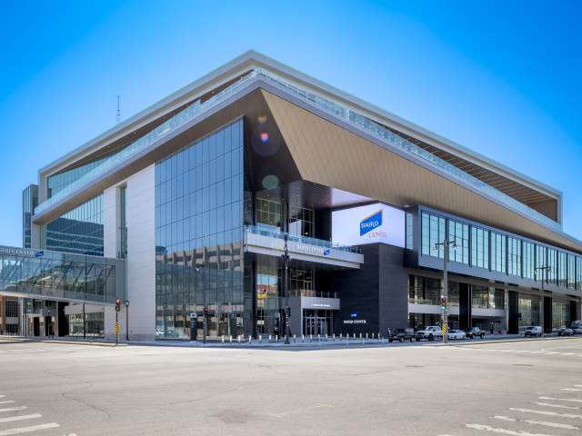 A daytime exterior view of the Baird Center in Milwaukee, showing its modern glass façade, angular roofline, and skywalk connection over the street, with cars and pedestrians nearby.
