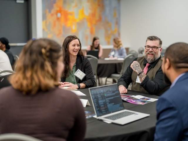 Attendees participate in a networking session around a round table at a conference. Several people smile and chat while reviewing materials and a laptop, creating a collaborative, professional atmosphere in a meeting room.