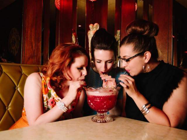 Three women sit together in a dimly lit lounge booth, sharing a large red cocktail served in a glass bowl and sipping it through straws. The setting has a retro, upscale feel with tufted seating, warm wood paneling, and moody lighting.