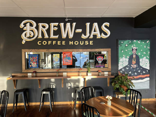 Interior of Brew-Jas Coffee House with a dark accent wall featuring the café’s name in large vintage-style lettering. A wooden counter with barstools sits beneath colorful framed artwork, and a vibrant painting of a traditionally dressed figure is displayed to the right. Sunlight streams across wooden tables and black chairs in the foreground.