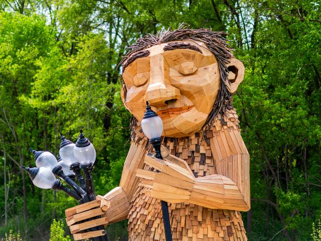 A large wooden sculpture of 'Mama Rosa,' a Thomas Dambo troll installation in Wauwatosa. The friendly giant figure is constructed from reclaimed wood pieces in natural tones, featuring a carved face with a gentle smile and twig-like hair made from bundled branches. The troll holds a bouquet of old-fashioned street lamps in one hand while lifting one lamp to her nose. The sculpture is set against a lush green forest backdrop with dense tree canopy, creating a whimsical scene that blends art with nature.