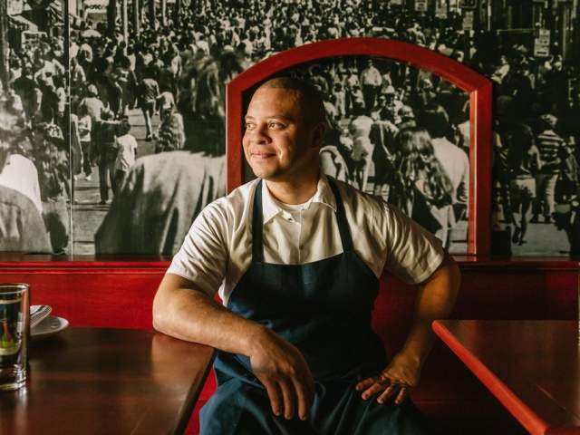 A man wearing a white shirt and dark apron sits at a restaurant table with his hands resting on the surface. He looks off to the side with a slight smile. The booth and table are dark red, and a large black and white mural of a crowded city street fills the background.