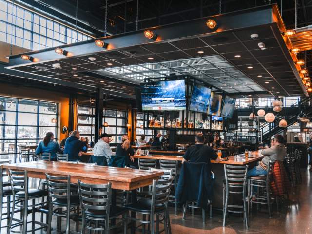 Guests sit around a large bar inside The Explorium Brewpub, with sports playing on overhead TVs.