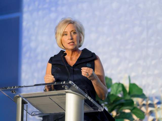 A woman with short blonde hair speaks at a clear podium with a microphone, wearing a sleeveless dark dress. She gestures with her hands while addressing an audience in a well-lit room with a patterned wall and greenery in the background.