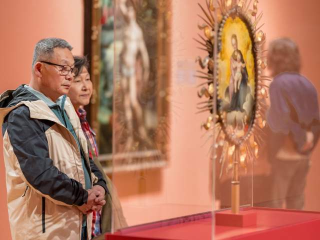 A middle-aged man wearing glasses and a tan-and-black jacket stands with his hands clasped, looking thoughtfully at an artwork in what appears to be an art museum or gallery. Behind him, a woman stands slightly out of focus, also observing the exhibit.

In the foreground on the right, there is a blurred religious-style artwork or icon displayed in a glass case, with a gold decorative frame. The overall scene has warm lighting and a calm, contemplative mood.