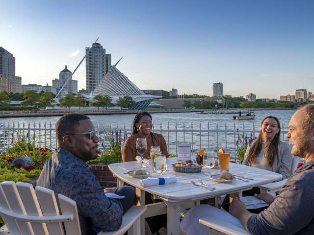 Four friends dining outdoors at Harbor House in Milwaukee, enjoying drinks and seafood by Lake Michigan with the Milwaukee Art Museum and city skyline in the background.
