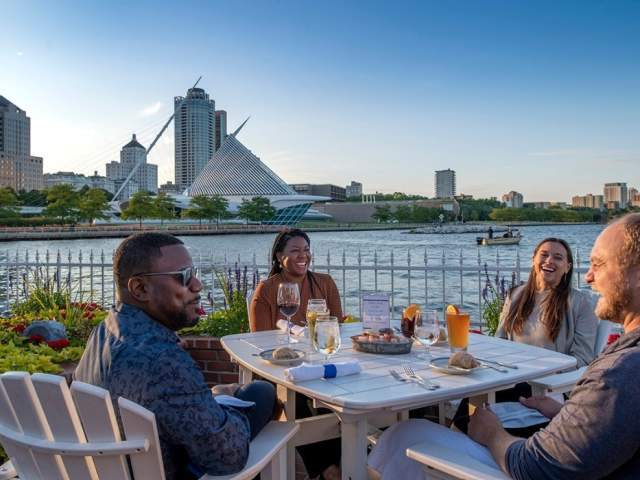 “Four friends dining outdoors at Harbor House in Milwaukee, enjoying drinks and seafood by Lake Michigan with the Milwaukee Art Museum and city skyline in the background.