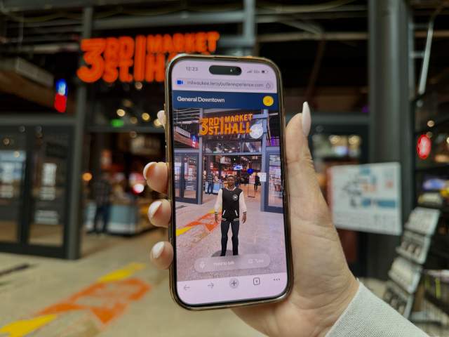 A person holds up a smartphone displaying an interactive digital guide inside 3rd Street Market Hall, with the venue entrance visible in the background.