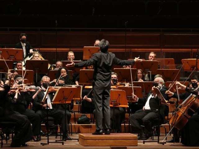A conductor stands on a podium with arms outstretched, leading a full orchestra onstage as musicians play string and brass instruments in a concert hall.