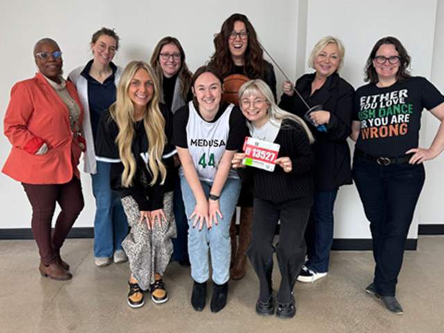 Group photo of nine women standing and crouching together indoors against a plain wall, smiling at the camera. One person in the front holds up a race bib while another wears a green sports jersey with the number 44. The group is dressed in a mix of casual and athletic clothing.