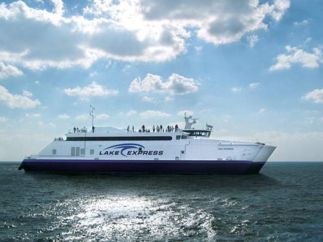 The Lake Express high-speed ferry crossing Lake Michigan on a sunny day, with passengers standing on the upper deck under a partly cloudy sky.