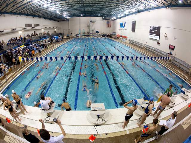 Wide overhead view of a large indoor swimming pool during a swim meet, with swimmers competing in multiple lanes while spectators and coaches watch from the pool deck and bleachers.