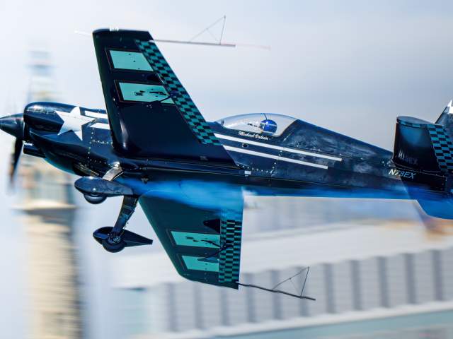 A blue aerobatic airplane performs a low-altitude maneuver over water, creating a mirrored reflection on the calm surface below. Motion blur in the background emphasizes the aircraft’s speed and precision during an air show performance.