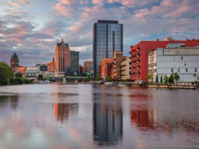 A view of the Milwaukee River at sunset with calm water reflecting the city skyline. Historic brick buildings line the right riverbank, while modern high-rises and iconic downtown towers rise in the background under a sky filled with soft pink and blue clouds.
