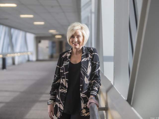 A woman with short blond hair stands smiling in a bright skywalk corridor, resting one hand on the railing. She wears a patterned blazer over a black top and is lit by natural daylight from the large windows beside her.